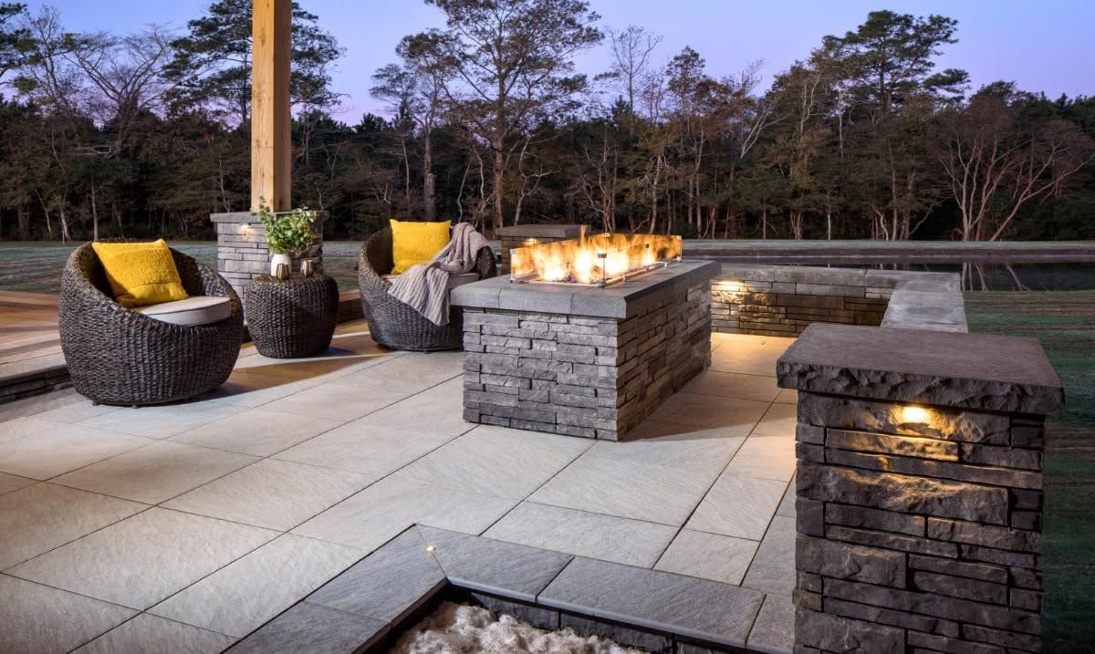 Outdoor patio with wicker chairs around a stone fire pit and a lit brick pillar at dusk