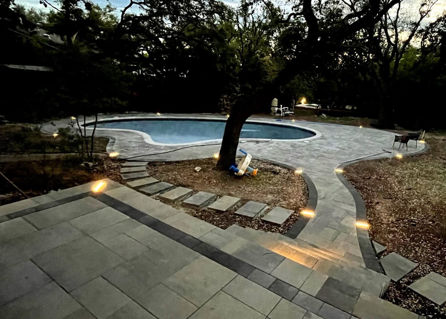 Curved stone patio leading to a lit backyard swimming pool at dusk, surrounded by trees and gravel paths