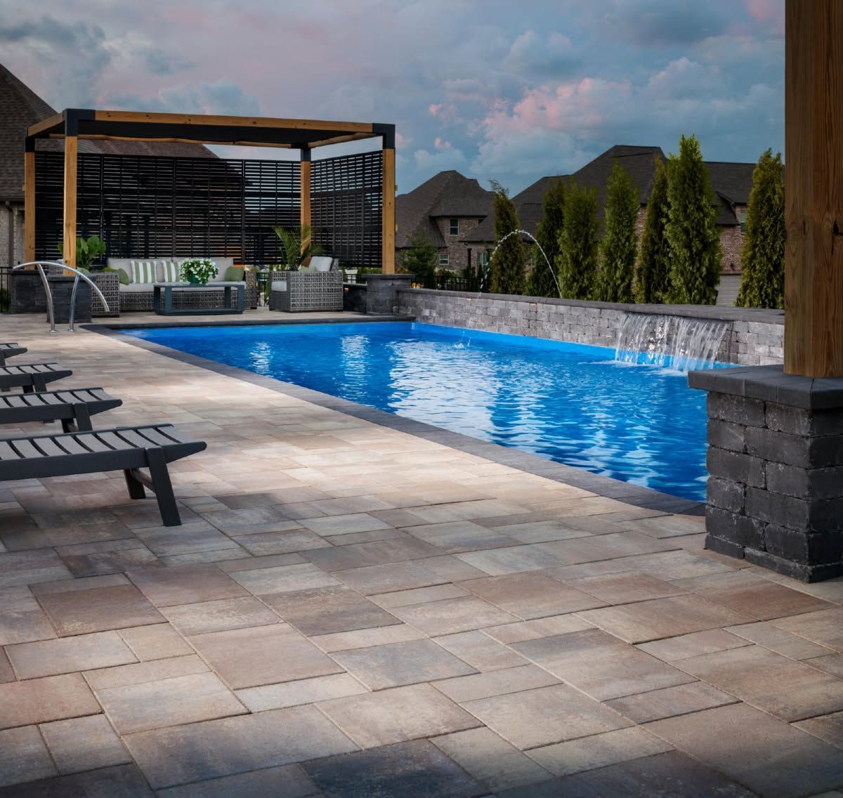 Backyard pool with blue water, stone patio, lounge chairs, and pergola at dusk