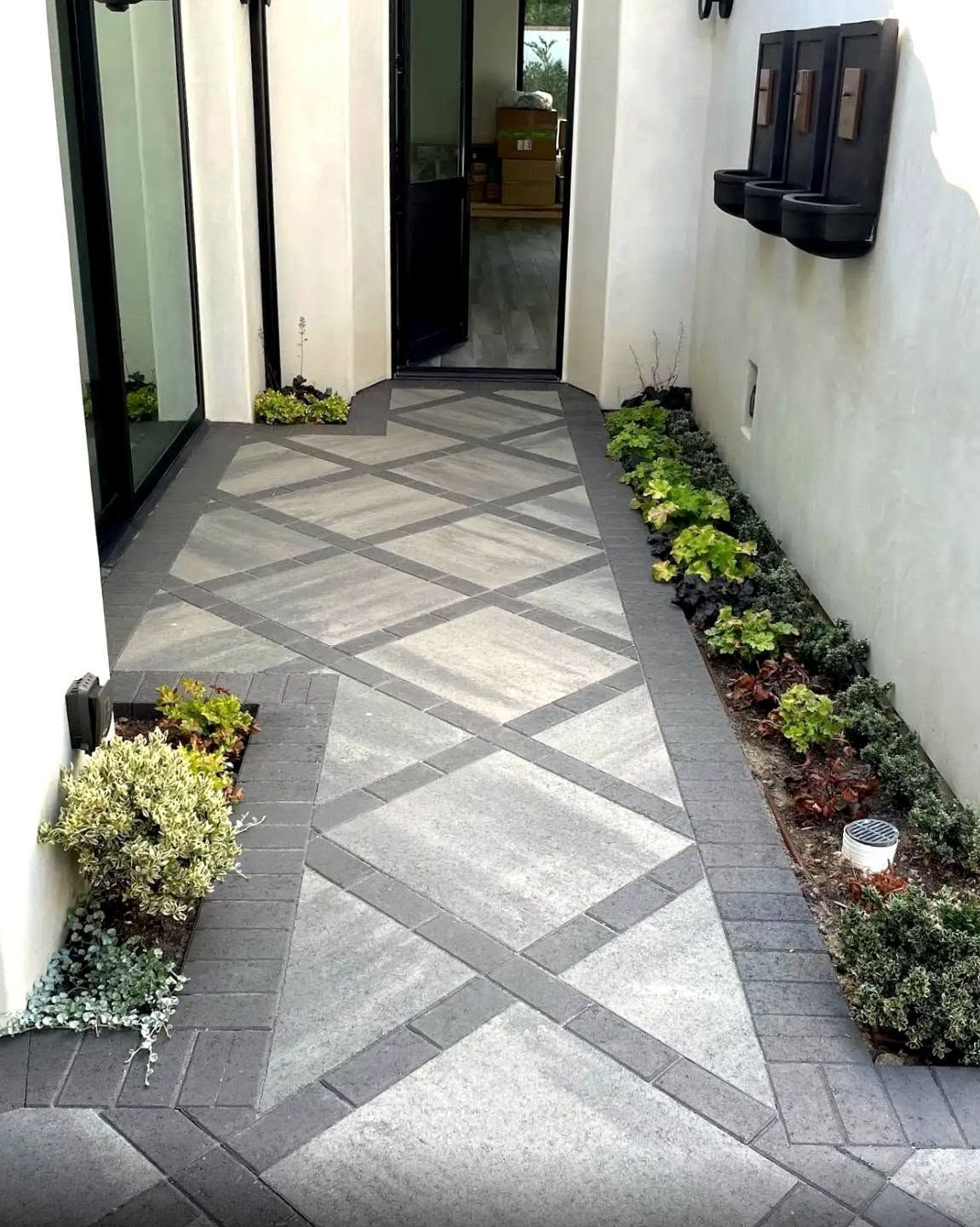 Narrow tiled walkway with geometric gray pattern, bordered by small plants and white walls.