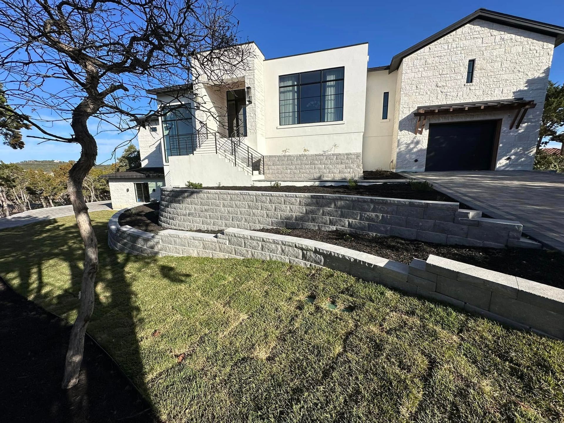 Modern stone house with dark garage, sloped driveway, and grassy front yard under a tree