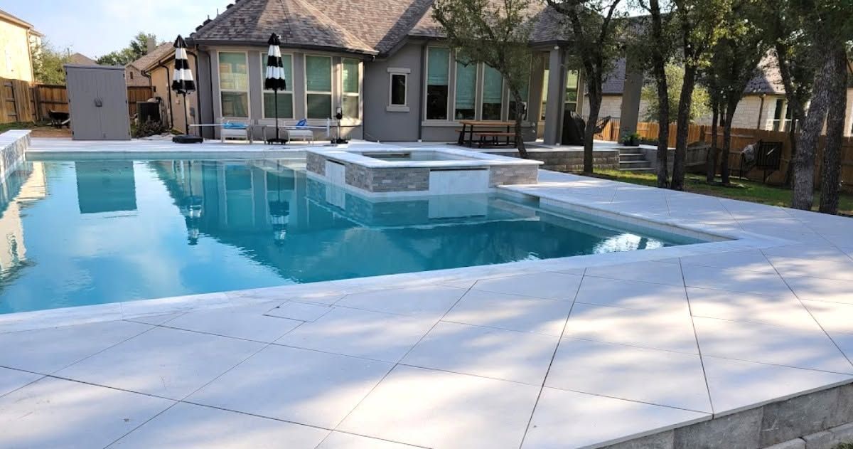 Modern backyard pool with clear blue water, stone patio, and a house with a tiled roof in the background