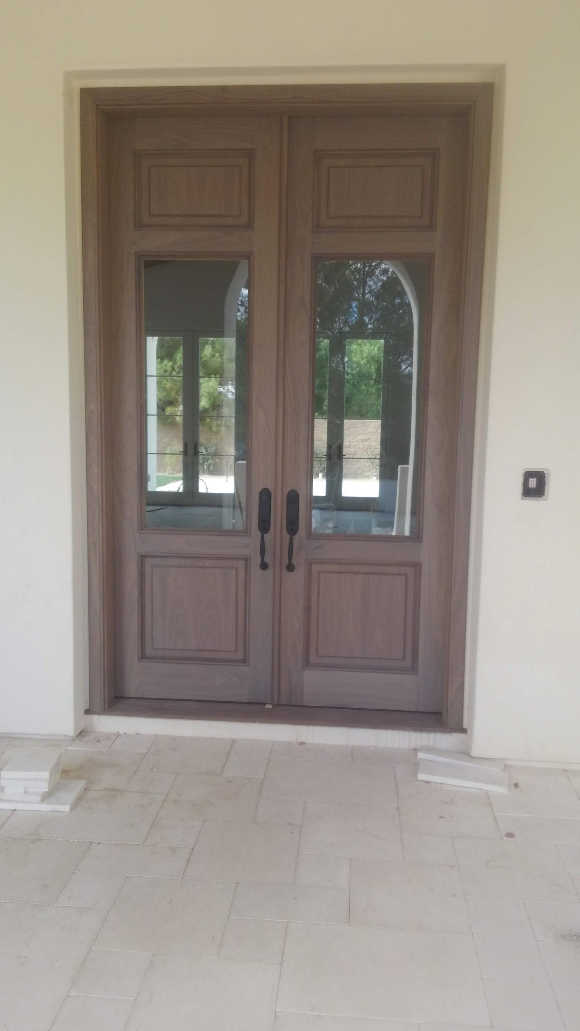 Double wooden doors with glass panels, brown frame, stone patio, white wall.