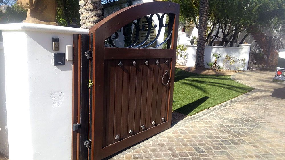 Wooden gate, partially open, with ironwork detail, set in a paved driveway, with a white pillar to the left.