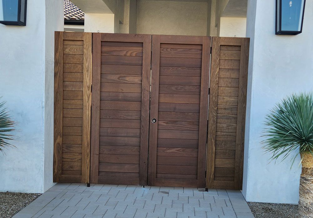 Wooden gate on a light-colored building. Dark brown gate is closed, flanked by side panels, on a paved walkway.