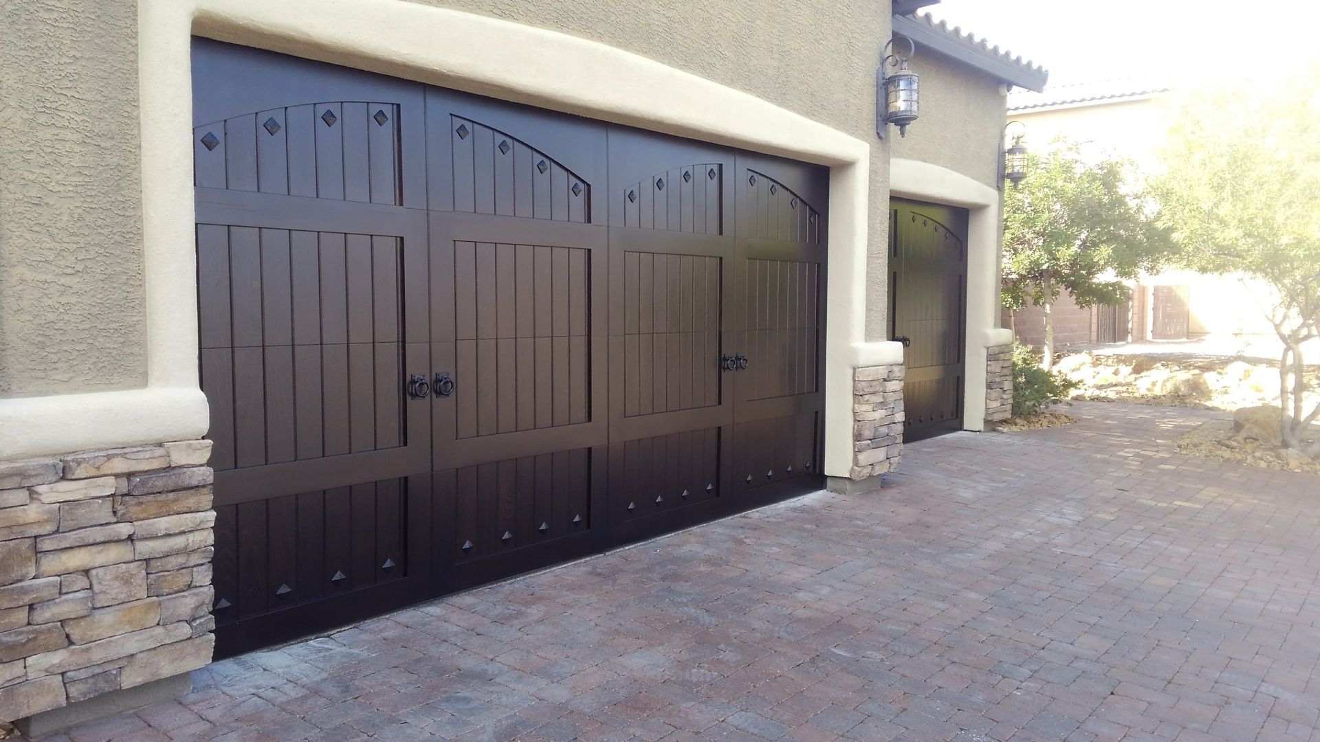 Wooden double gate with arched top, set between white pillars and a black fence, on a paved driveway.