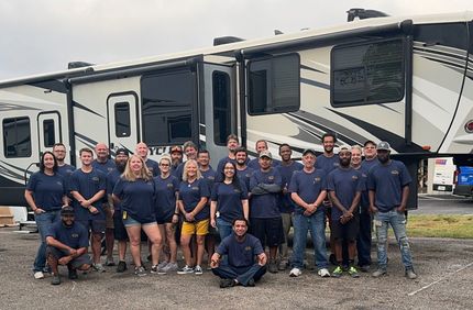 Group of people in blue shirts pose in front of a large RV in an outdoor setting.