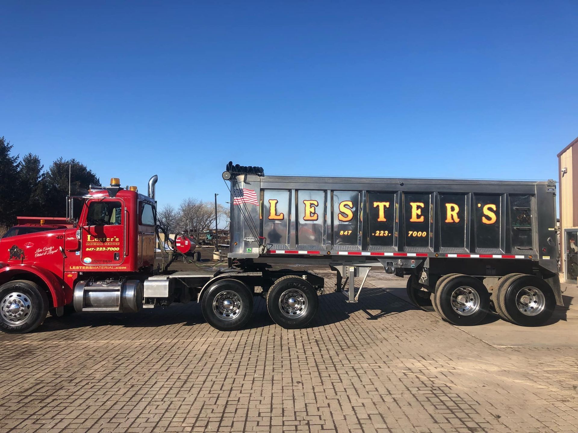 Red and black Lester's dump truck parked on brick, under a bright blue sky.