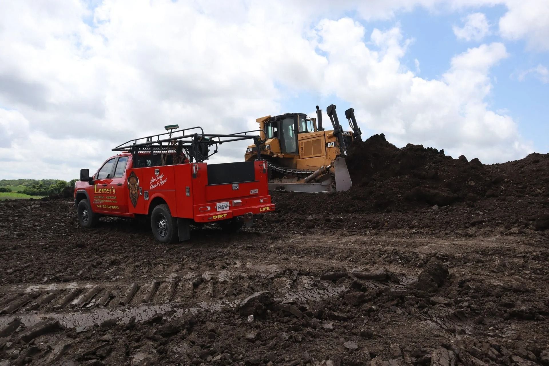 Red truck and bulldozer working on a pile of dark soil under a cloudy sky.