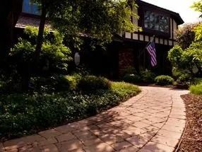Brick pathway leading to a house with a flag; surrounded by green bushes and trees.