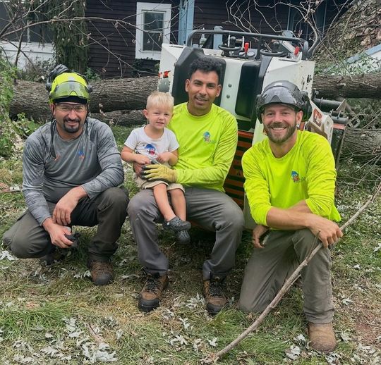 Three workers in hard hats crouch with a toddler outdoors in front of a utility truck.