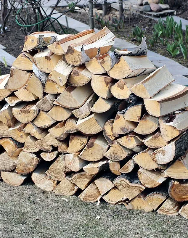 Pile of cut firewood stacked outdoors on grass and pavement.
