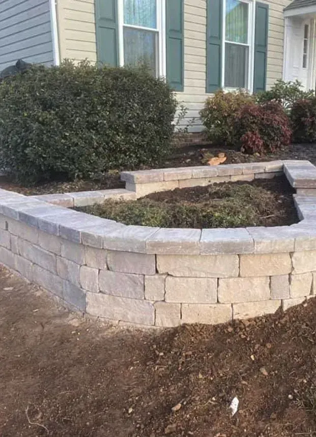 Brick patio with retaining wall and steps under a wooden structure, in a yard with brown grass.