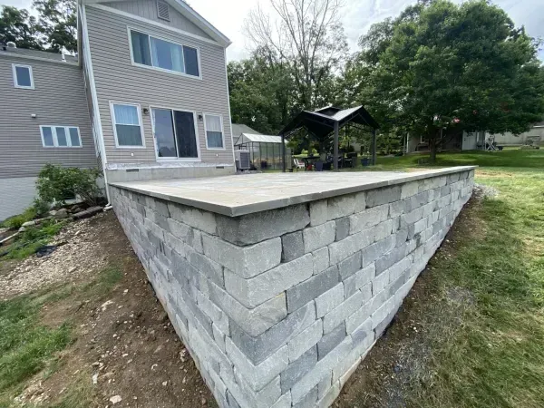 Gray retaining wall supports a concrete patio behind a two-story house with a sliding glass door.