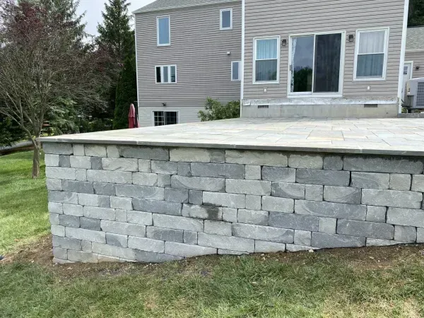 Stone retaining wall supporting a paved patio, with a gray house in the background.