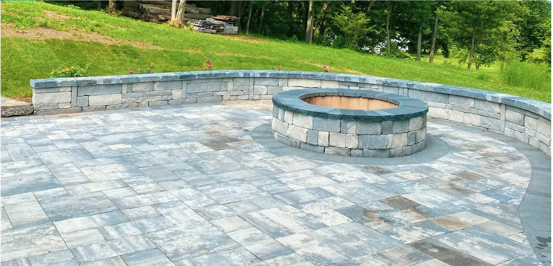 Stone fire pit and patio surrounded by a curved retaining wall and green grass.