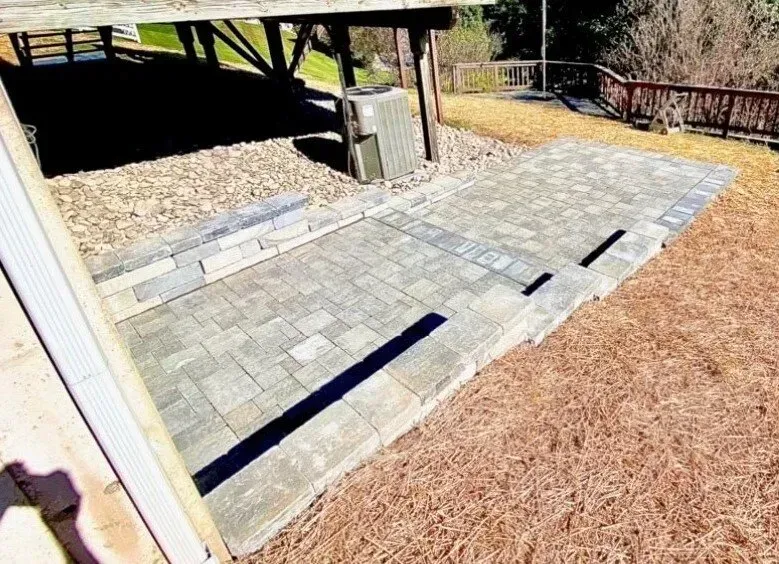 Brick patio with retaining wall and steps under a wooden structure, in a yard with brown grass.