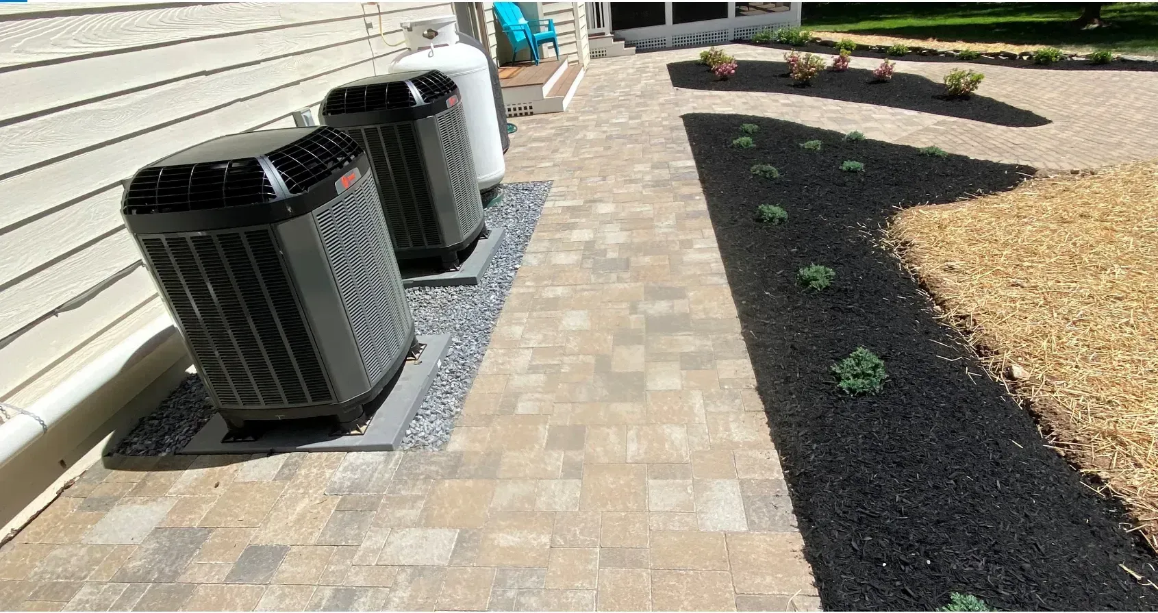 Two black HVAC units and a propane tank on pavers next to a gravel bed.