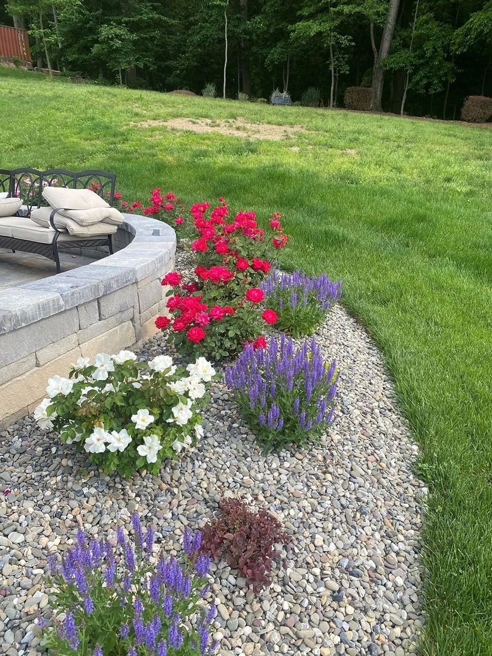 Flowers in shades of red, white, and purple border a stone retaining wall with seating; a grassy lawn extends beyond.