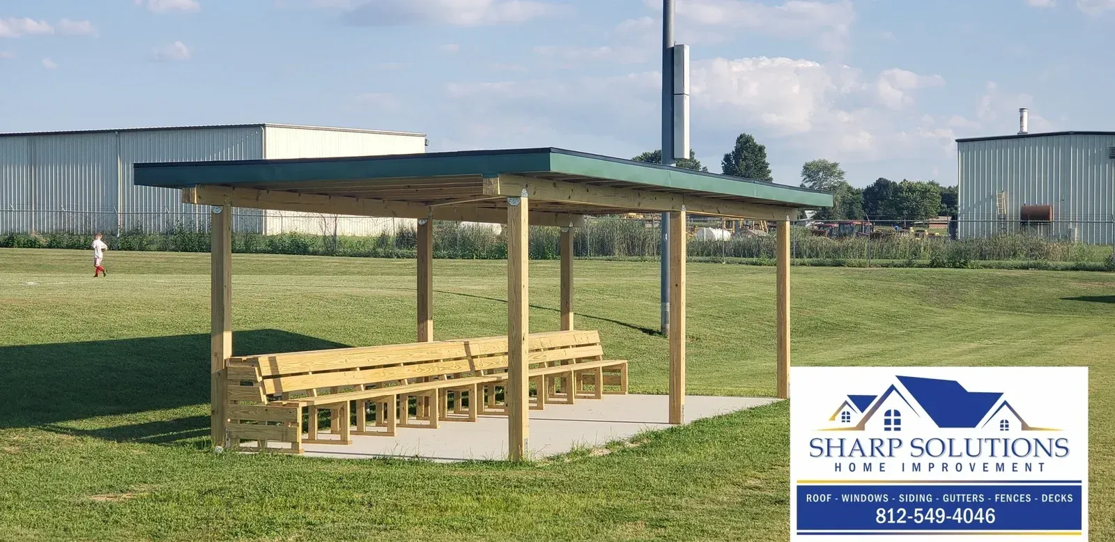 Outdoor pavilion with wooden benches on a grassy field, near industrial tanks under a partly cloudy sky