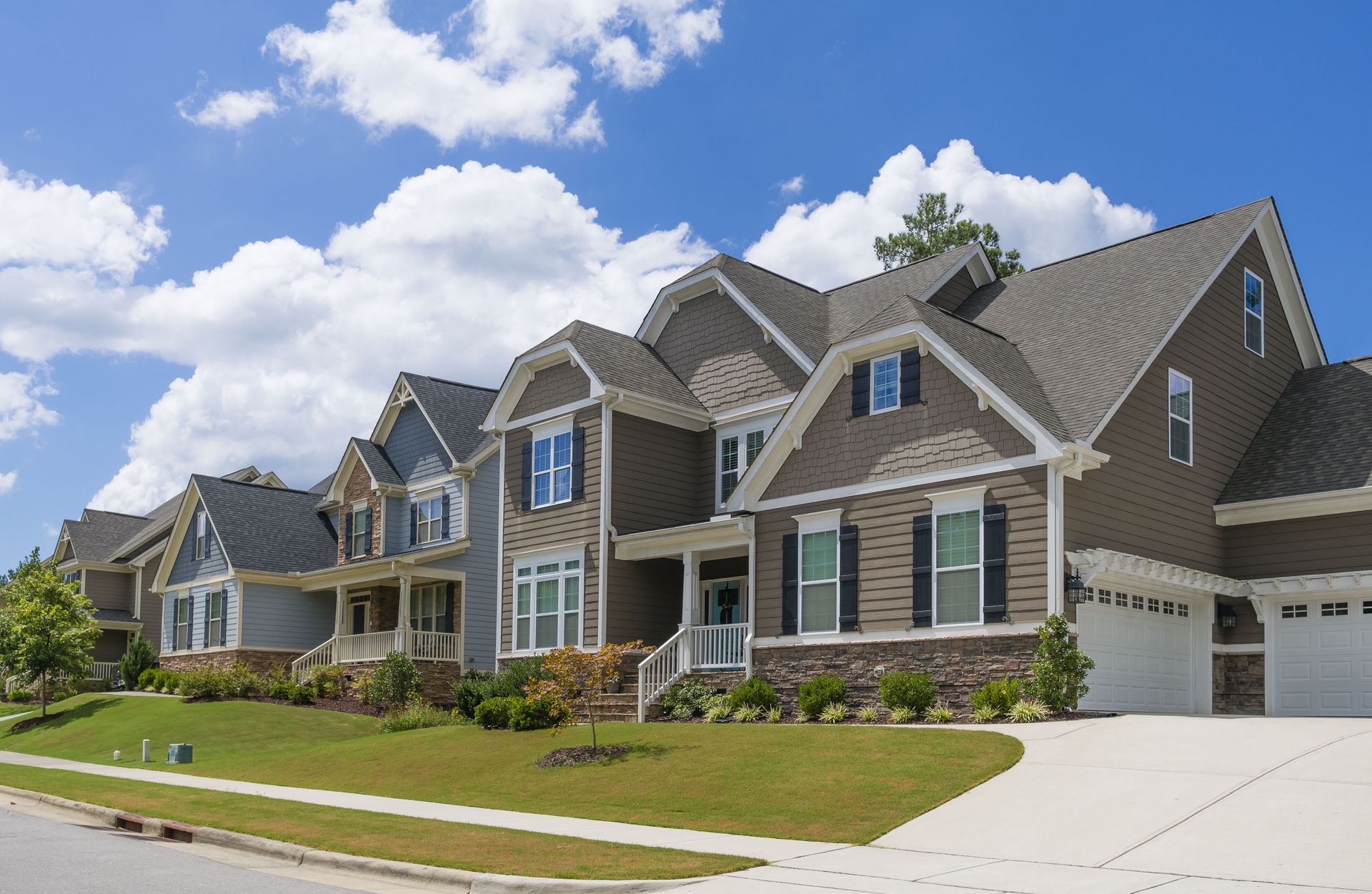 Large suburban brick houses with gabled roofs, a driveway, and a blue sky with clouds