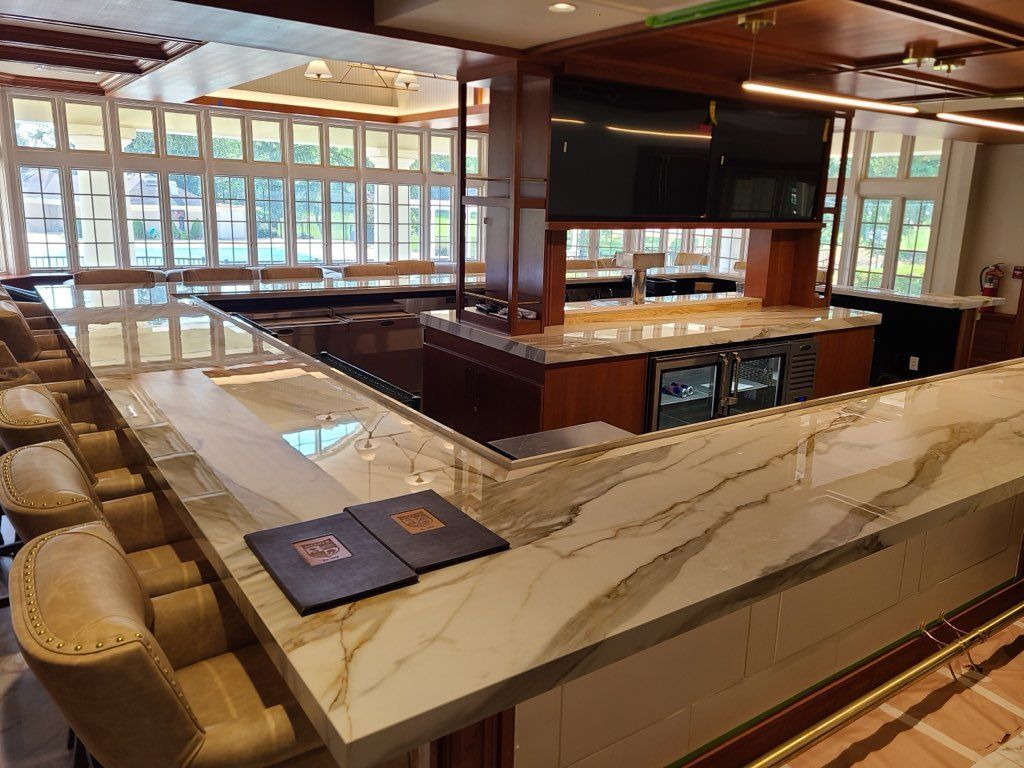 A bar with marble countertop, bar stools, and televisions, inside a room with large windows.