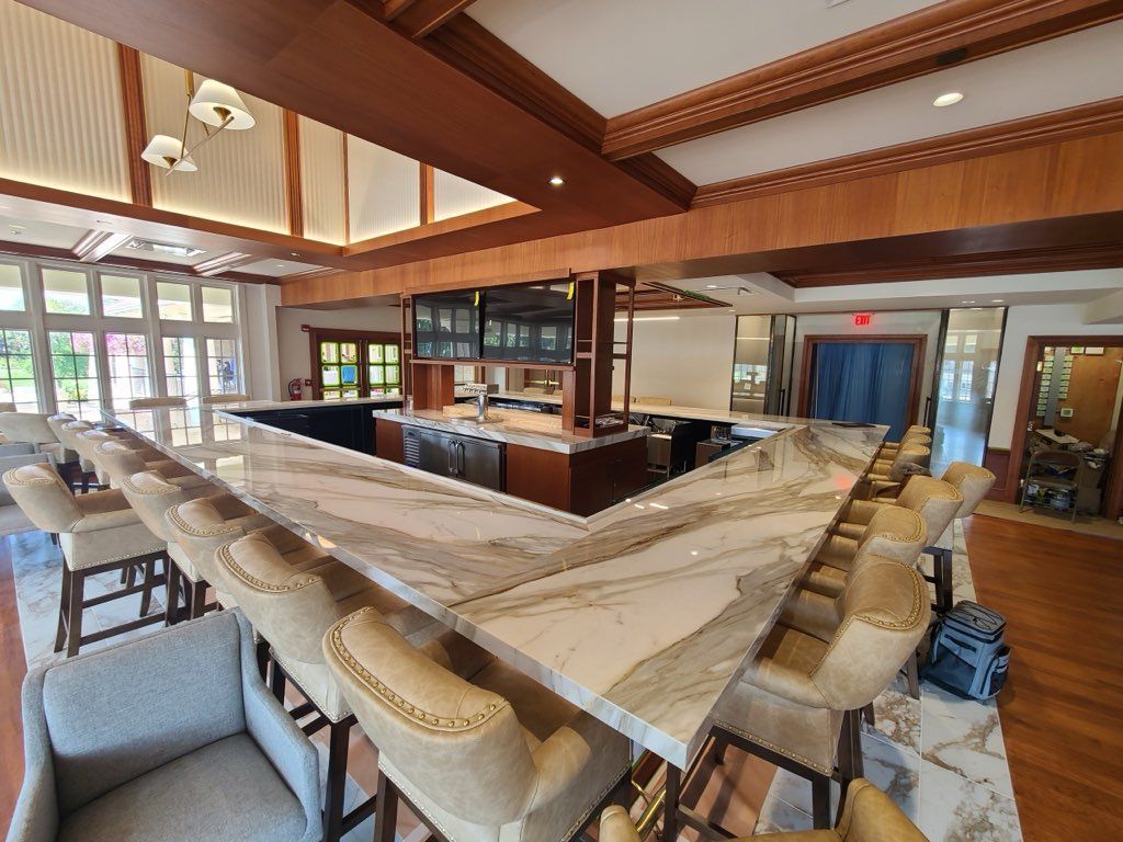 Bar with marble countertop and beige stools. Wooden accents, TV screens, and outdoor views through windows.