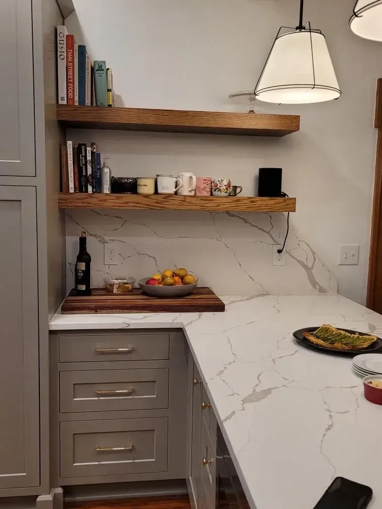 A kitchen with a marble counter top, wooden shelves, and a cutting board.