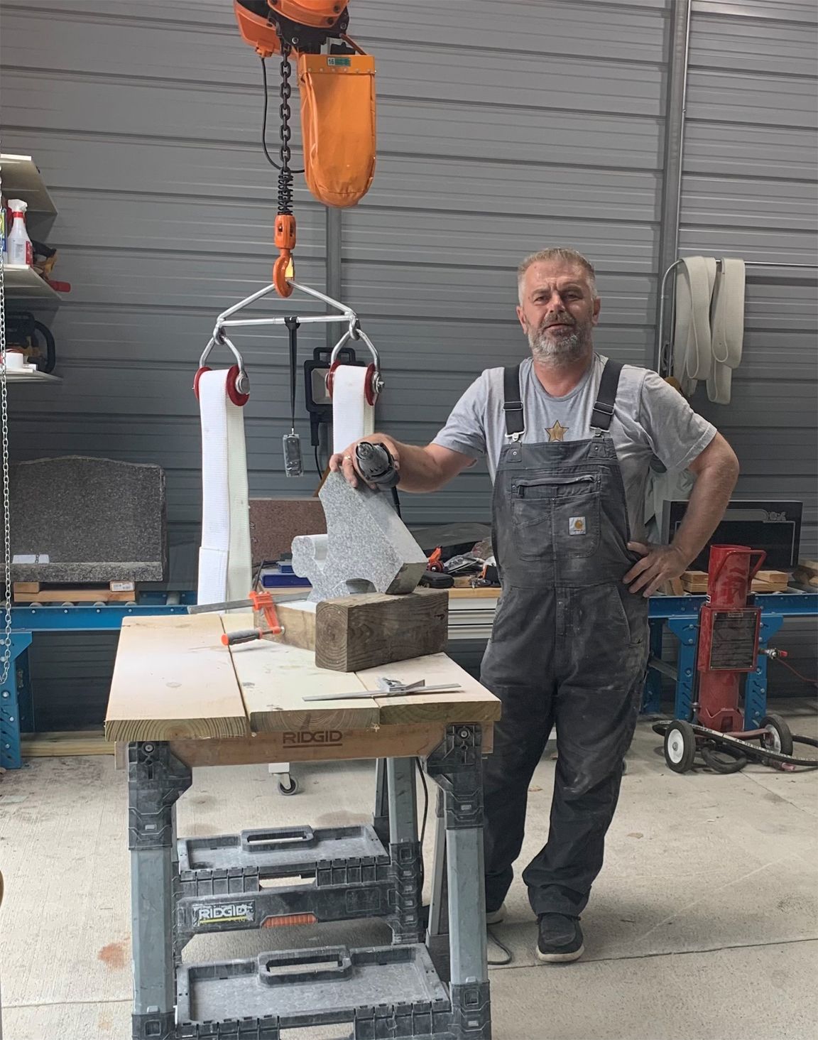 Sculptor in overalls stands near a work table with a stone sculpture and a lifting device in a workshop.