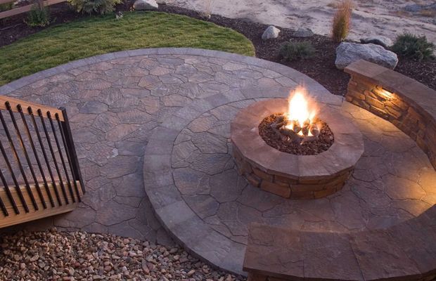 Stone patio with fire pit, built-in curved bench, and surrounding landscaping; flames are burning.
