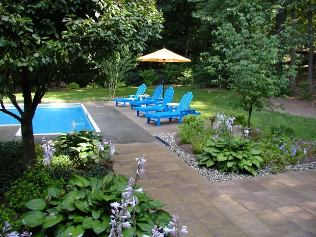 Blue lounge chairs sit by a pool, with an umbrella and garden in the background.