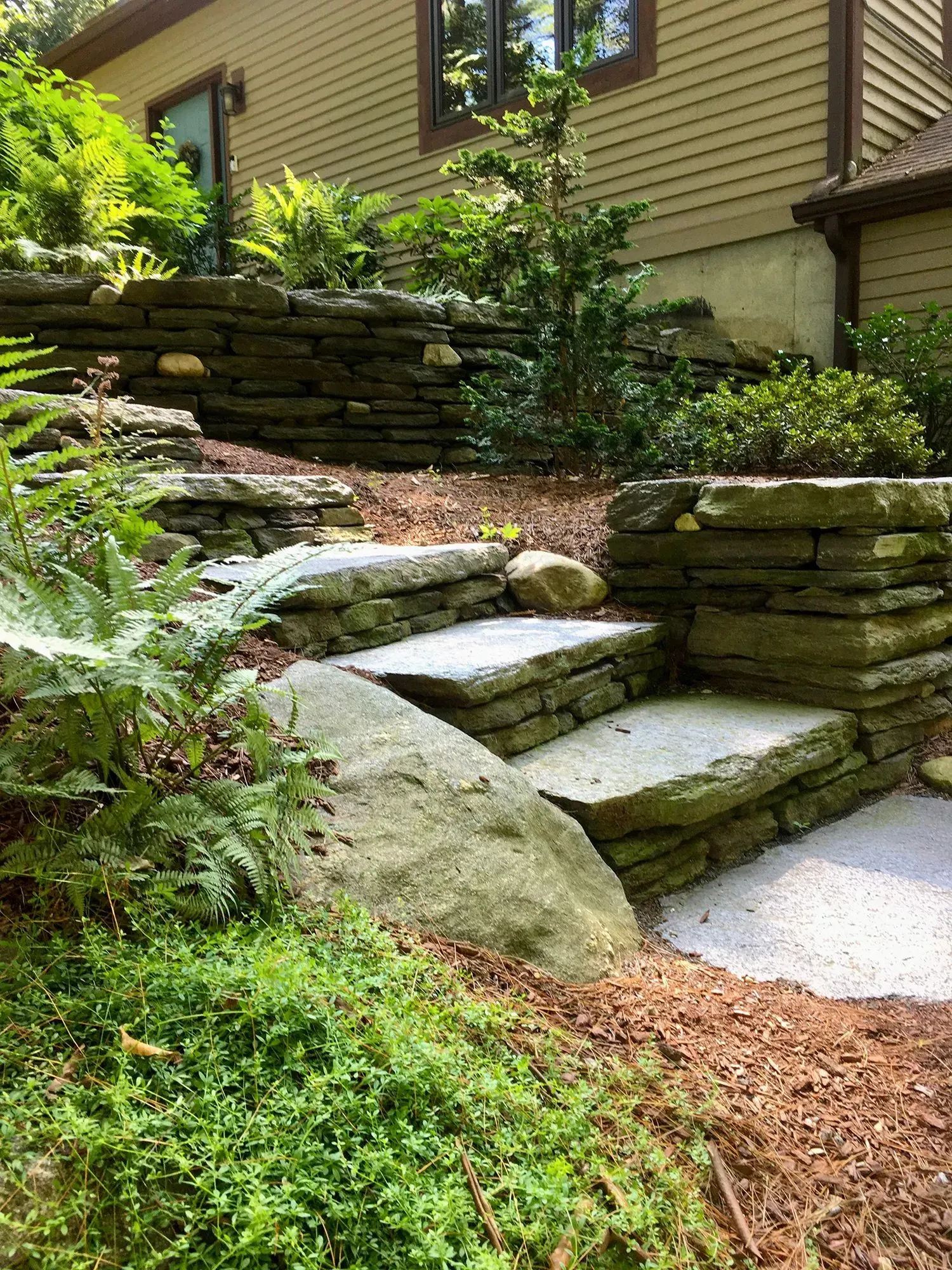 Stone steps and retaining walls on a hillside, leading to a house, surrounded by greenery.