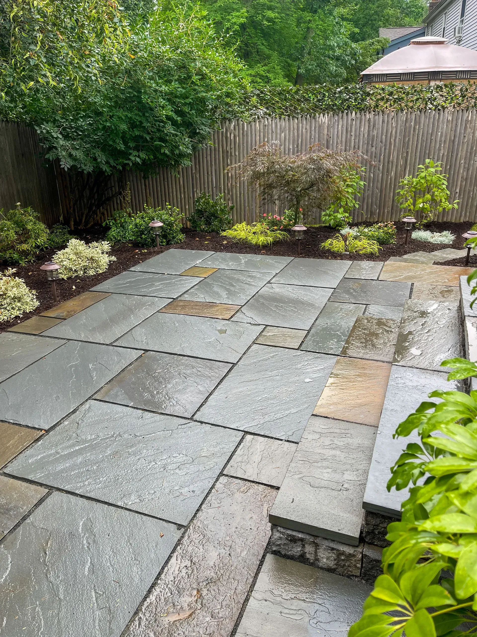 Stone patio with landscaping, including trees, plants, and a wooden fence in the background.