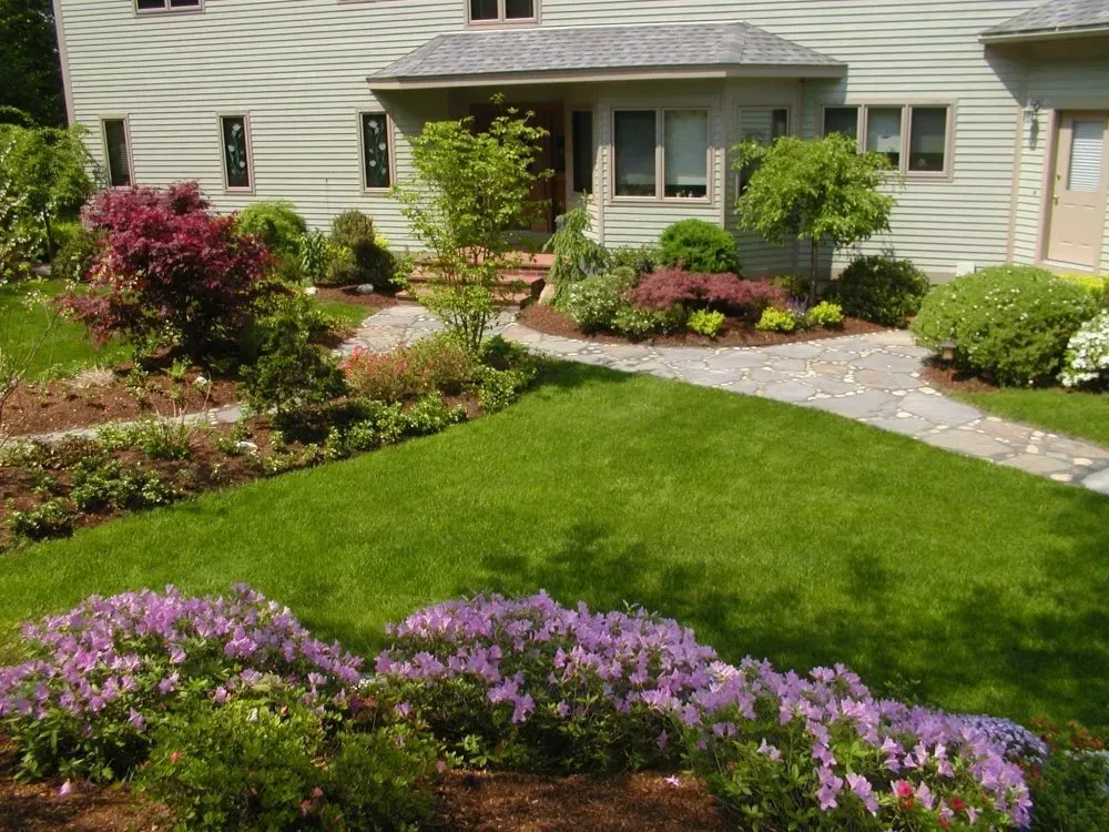 Lush green lawn with a stone pathway leading to a house. Colorful flowers and bushes line the lawn.