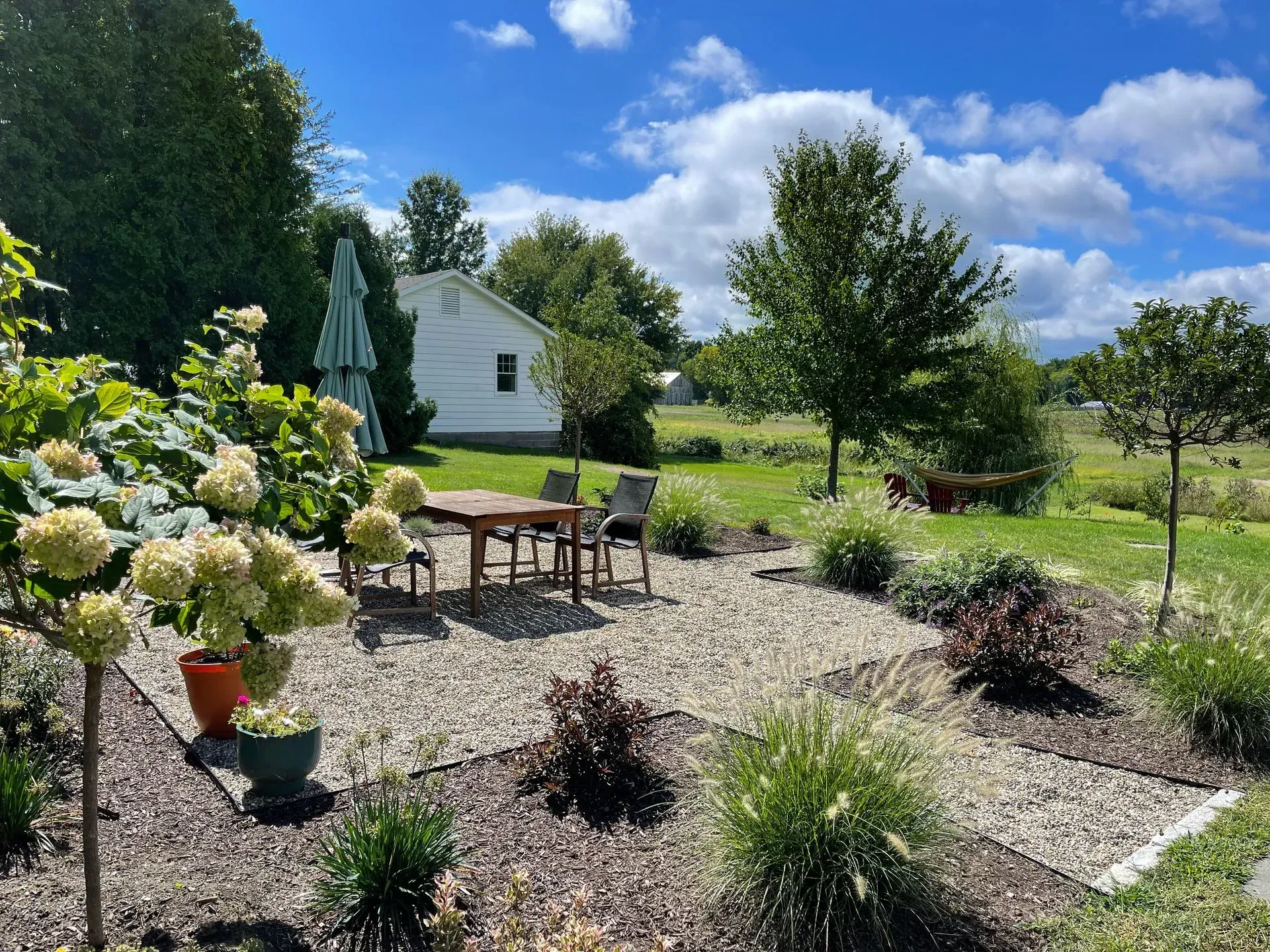 Patio with table and chairs in a landscaped garden, sunny day. White house in background.