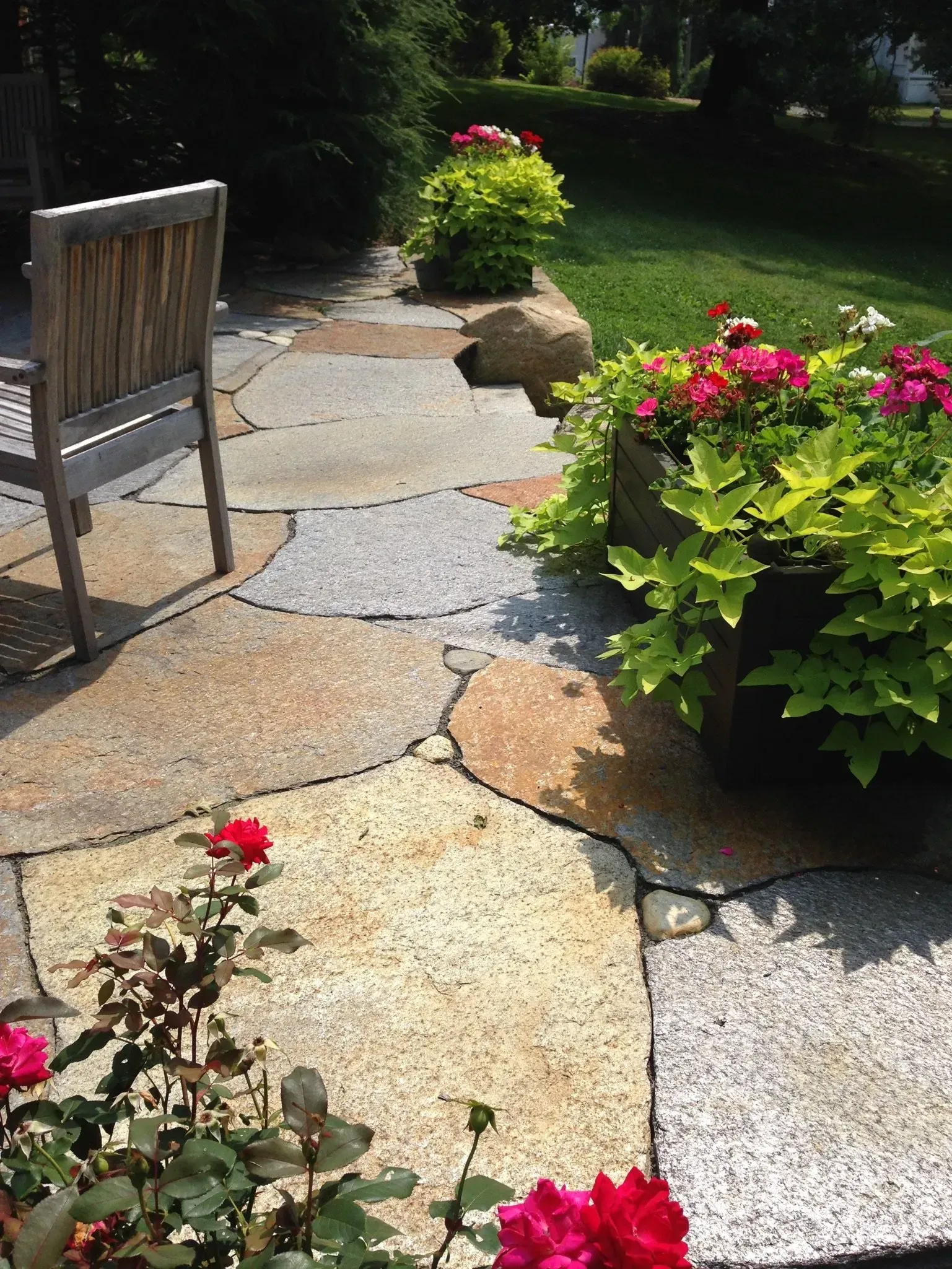 Stone patio with potted flowers, a wooden chair, and a grassy yard.