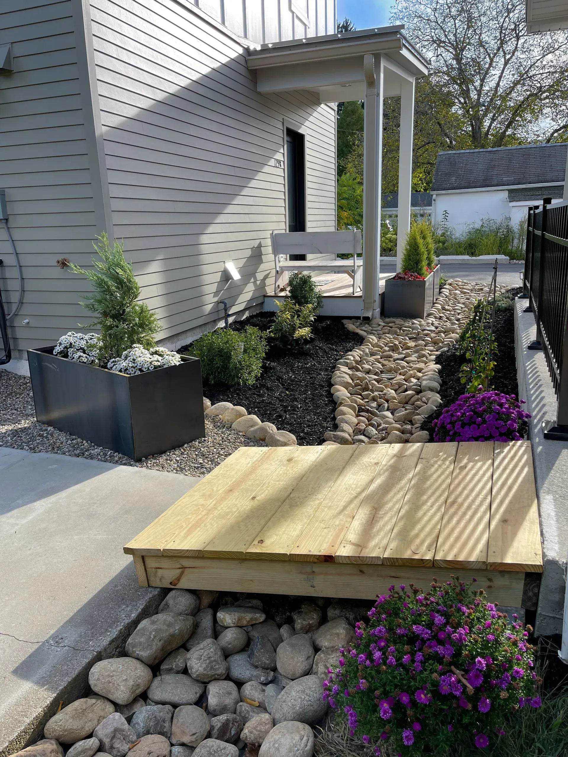 Wooden bridge over a rock-filled garden path leading to a porch with seating; plants and flowers surround.