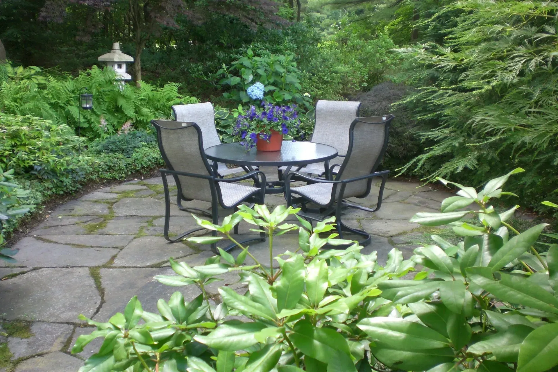 Outdoor patio with table, four chairs, and potted flowers, surrounded by greenery.