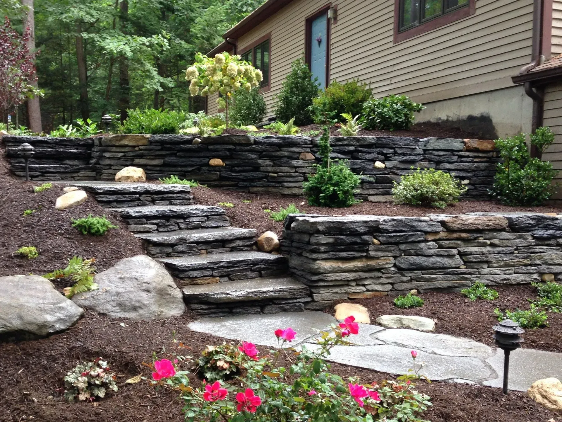 Stone steps and retaining walls with landscaping, including plants, mulch, and a house in the background.