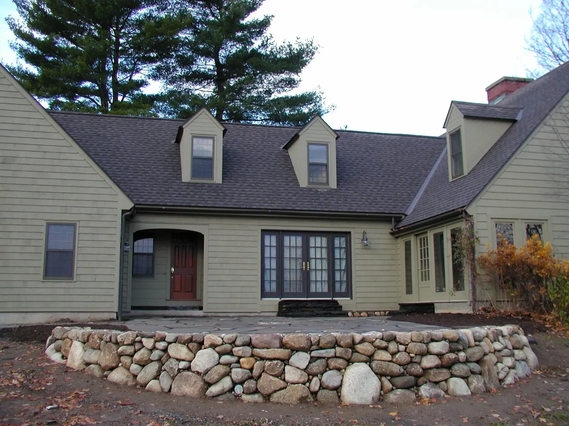 Stone patio in front of a light green house with dark roof and doors, trees in the background.