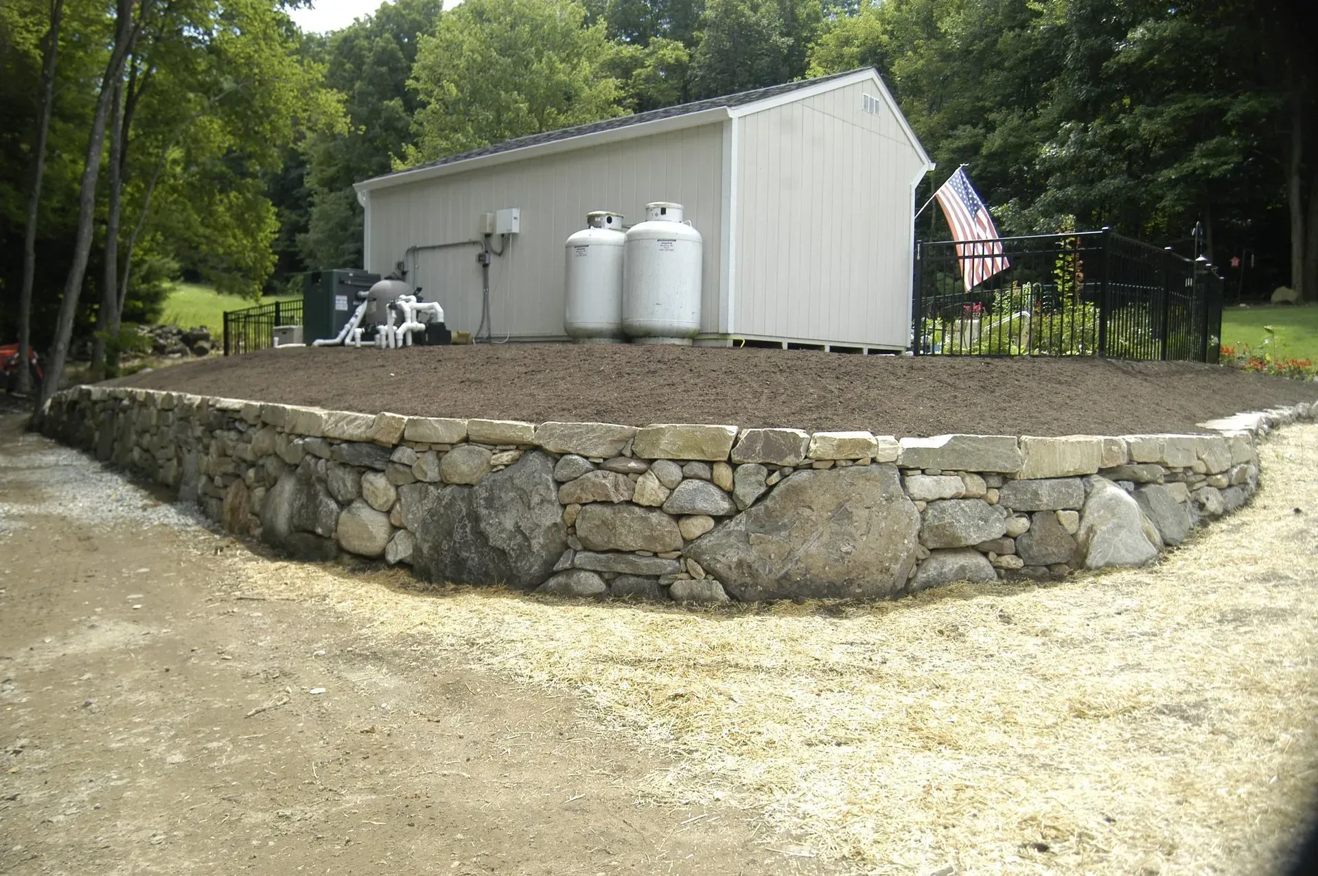 Stone retaining wall around a raised area with a white shed and propane tanks.