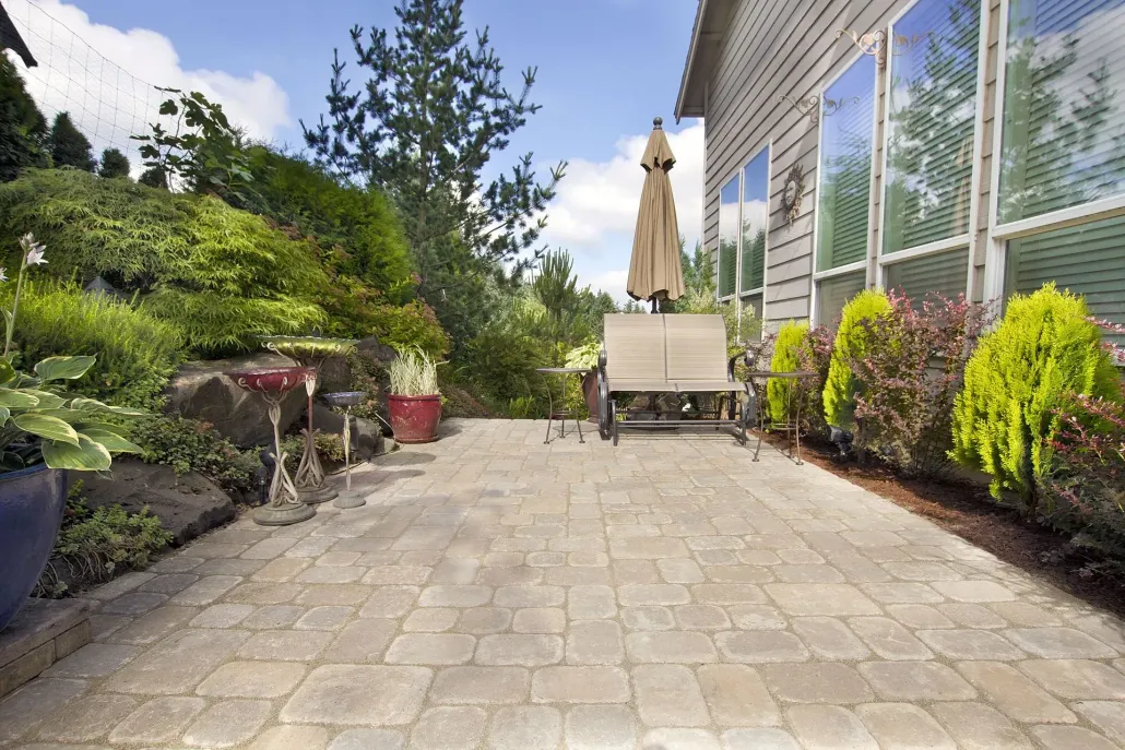 A stone patio beside a house featuring a bench, an umbrella, and surrounding lush green landscaping under a bright sky.