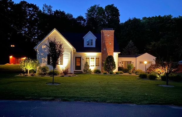 White house with lit exterior lights at dusk, green lawn, brick chimney, dark trees in background.