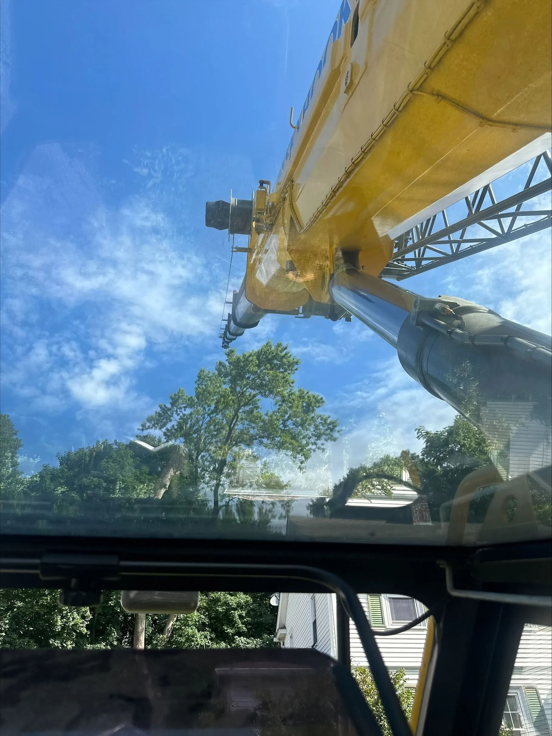 View from inside a yellow crane cab, looking up at the boom against a blue sky with trees.