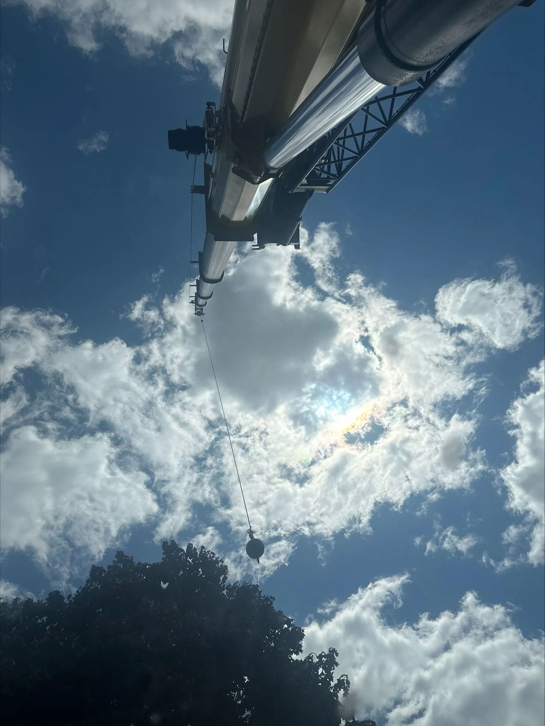 A crane arm against a bright blue sky with fluffy clouds; a dark, leafy tree in the lower left corner.
