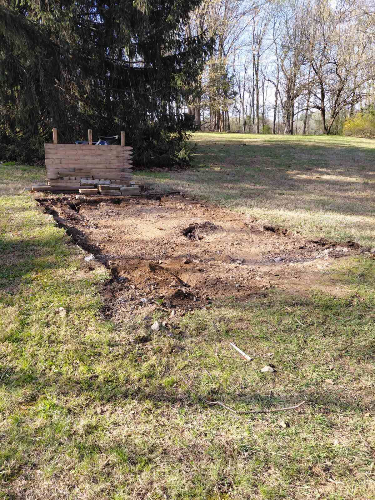 A cleared, rectangular dirt plot on a grassy lawn with a small wooden frame structure at the back.