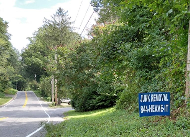 Roadside sign: Junk removal service advertisement. Blue sign with phone number, next to a rural road with trees.