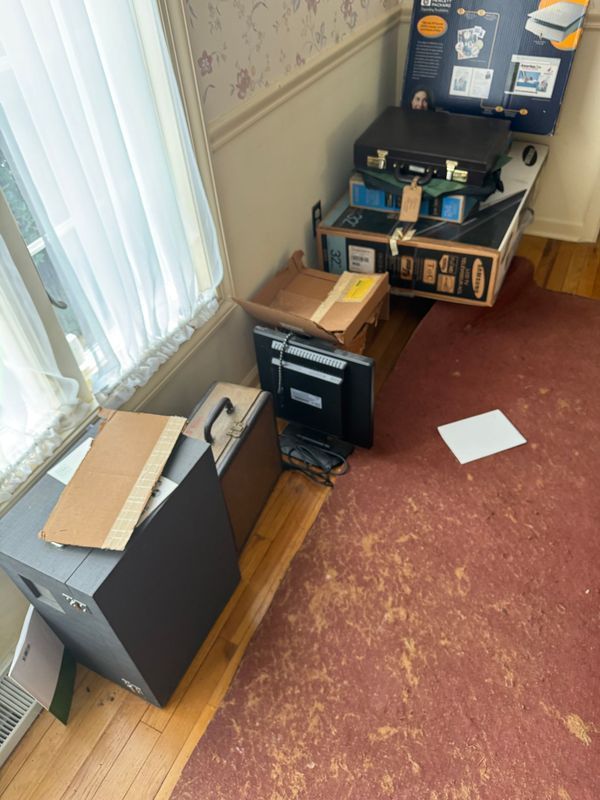 A pile of boxes, a computer, and a monitor in a room with a window and a red rug.