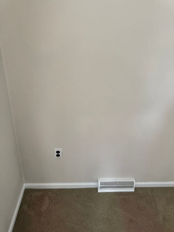 Beige interior wall with a white baseboard, outlet, and rectangular vent. Brown carpet.