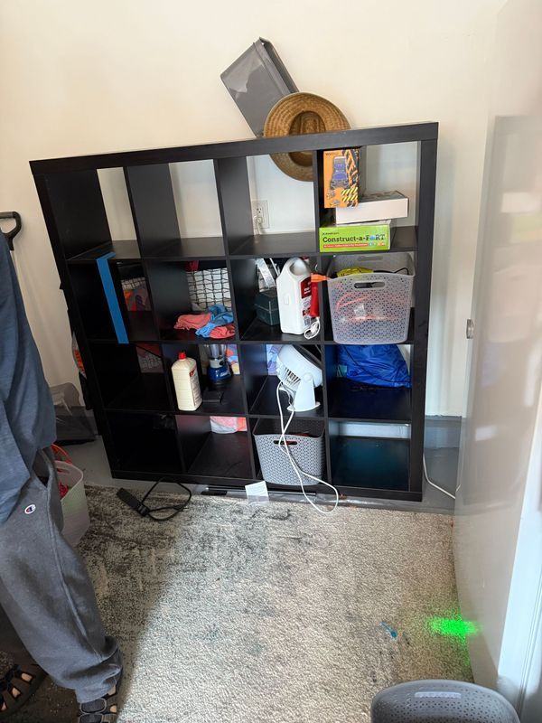 Black shelving unit with various items, against a white wall and light-colored carpet.
